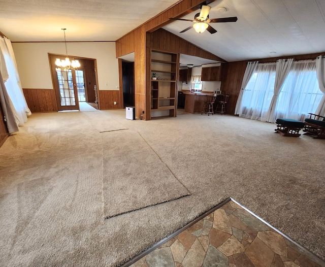 unfurnished living room featuring wood walls, wainscoting, lofted ceiling, ceiling fan with notable chandelier, and light colored carpet