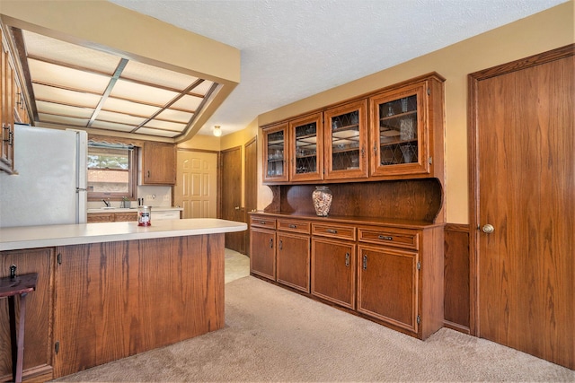 kitchen with glass insert cabinets, freestanding refrigerator, a peninsula, brown cabinets, and light colored carpet