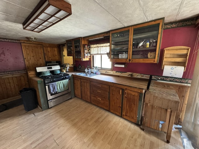 kitchen featuring stainless steel range with gas cooktop, glass insert cabinets, a sink, brown cabinets, and light wood finished floors