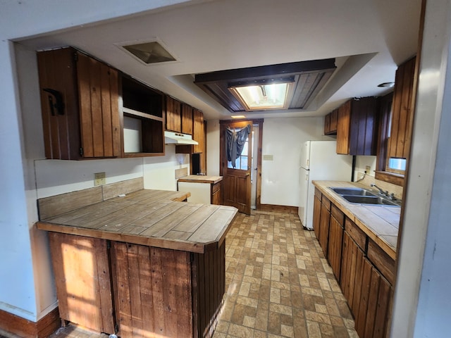 kitchen featuring freestanding refrigerator, brick patterned floor, under cabinet range hood, a sink, and a raised ceiling