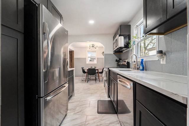 kitchen featuring marble finish floor, light stone counters, dark cabinets, arched walkways, and appliances with stainless steel finishes