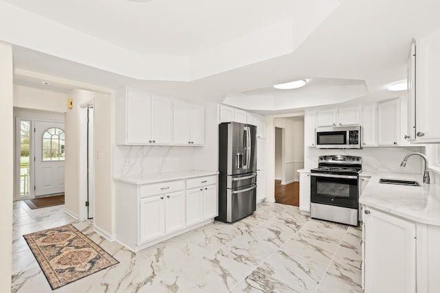 kitchen with appliances with stainless steel finishes, white cabinets, a raised ceiling, marble finish floor, and a sink