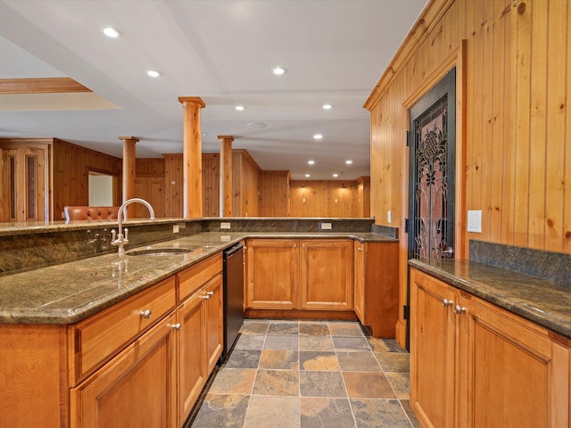 kitchen featuring dark stone counters, a sink, wood walls, dishwasher, and brown cabinetry
