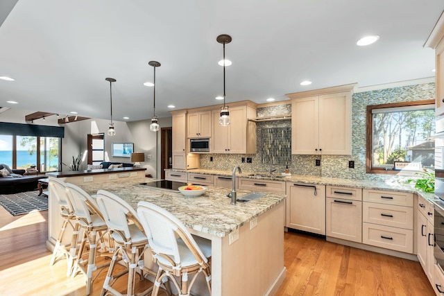 kitchen featuring stainless steel microwave, an island with sink, backsplash, light wood-style floors, and a sink