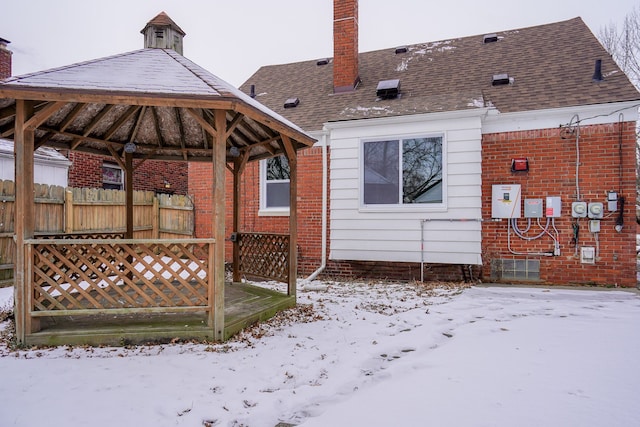 snow covered rear of property with a shingled roof, a gazebo, a chimney, fence, and brick siding