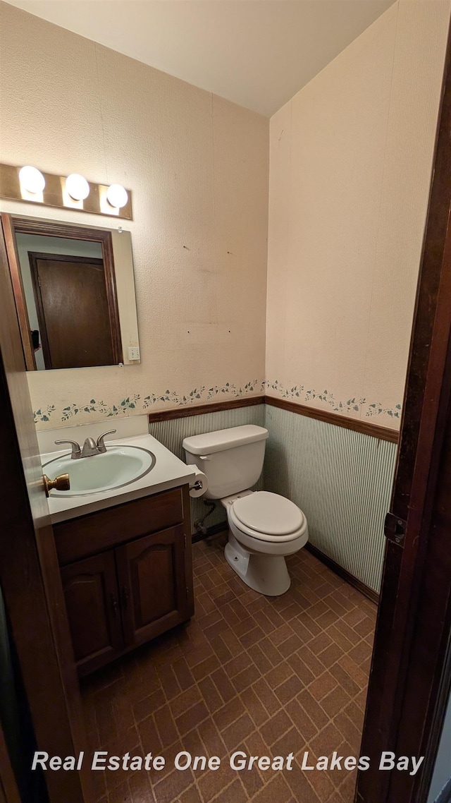 half bath with toilet, brick patterned floor, wainscoting, and vanity
