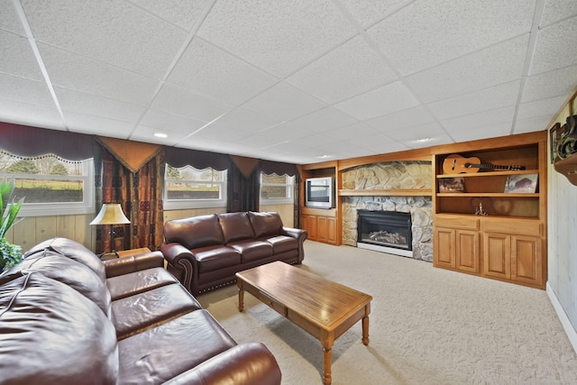 carpeted living area featuring wood walls, built in shelves, a stone fireplace, and a drop ceiling