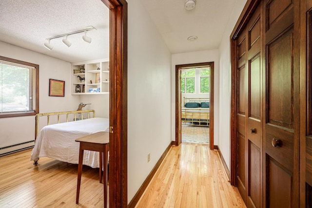 hallway featuring a baseboard radiator, a textured ceiling, light wood-style floors, rail lighting, and baseboards