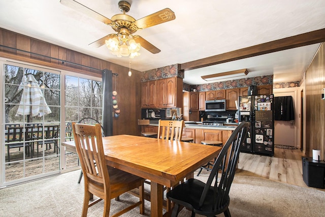 dining area with wooden walls, beam ceiling, ceiling fan, and light colored carpet