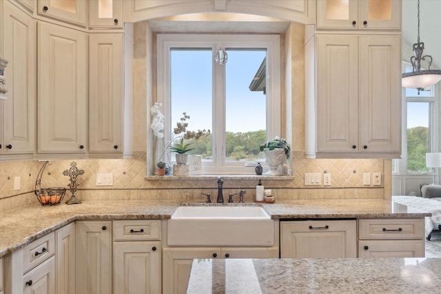 kitchen featuring a sink, glass insert cabinets, backsplash, light stone countertops, and hanging light fixtures