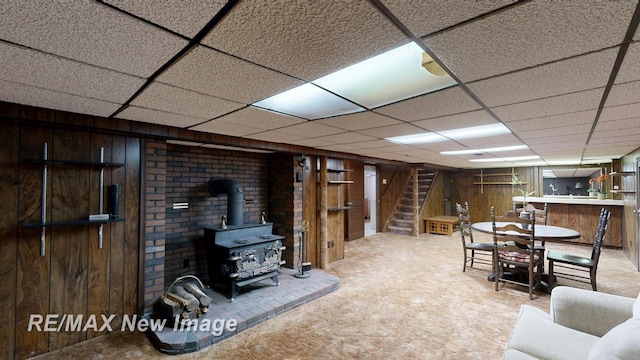 carpeted living area with a wood stove, bar area, stairway, wood walls, and a drop ceiling