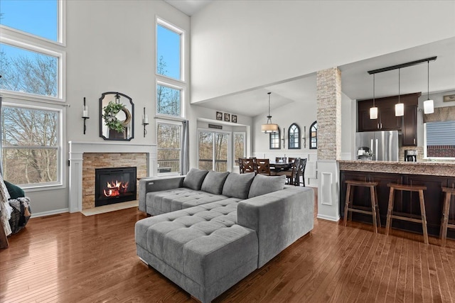 living room with a towering ceiling, a stone fireplace, a wealth of natural light, and dark wood finished floors