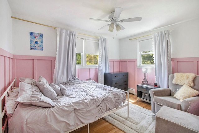 bedroom with a decorative wall, multiple windows, wainscoting, wood finished floors, and ceiling fan