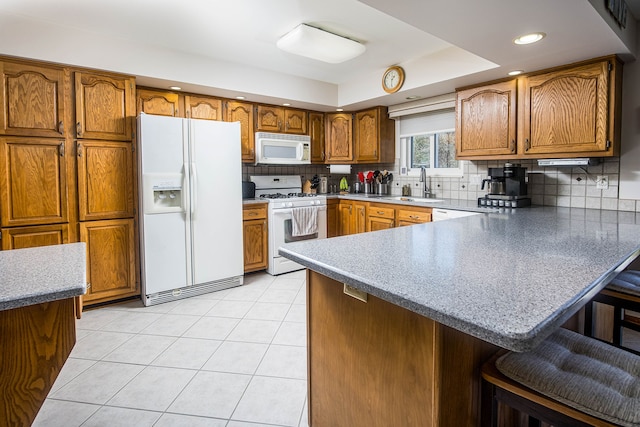 kitchen featuring a tray ceiling, a sink, white appliances, a peninsula, and brown cabinetry