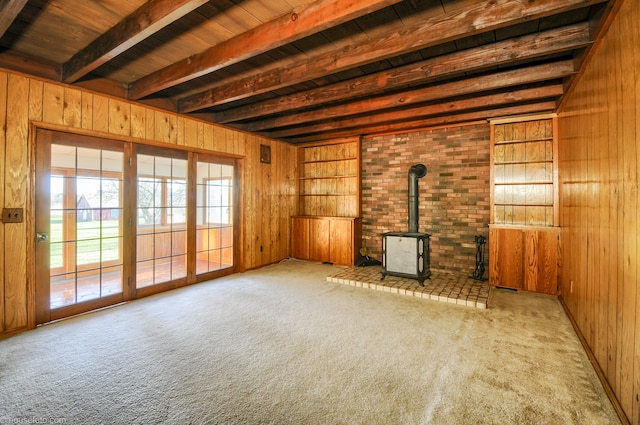 unfurnished living room featuring beamed ceiling, wooden walls, a wood stove, and carpet flooring