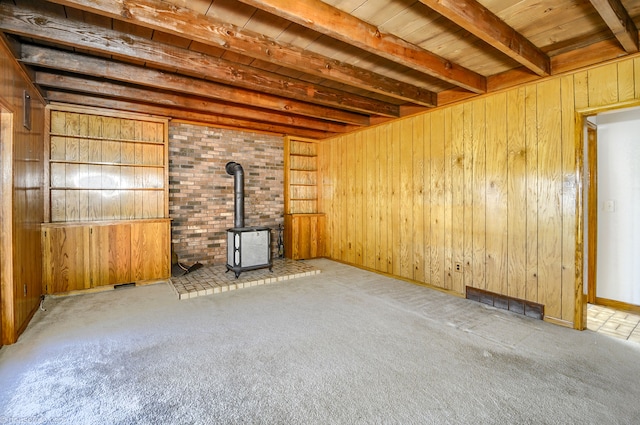 unfurnished living room featuring carpet floors, a wood stove, beamed ceiling, wood walls, and visible vents