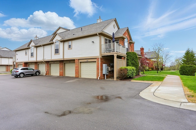 view of side of property with driveway, brick siding, a balcony, an attached garage, and a chimney