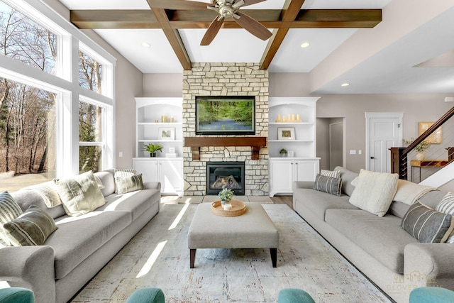 living room with a stone fireplace, light wood-type flooring, a ceiling fan, beamed ceiling, and stairs