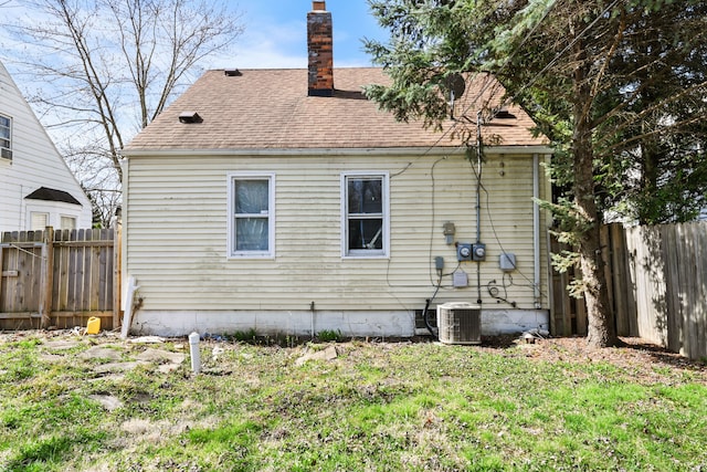 rear view of house with fence, roof with shingles, central AC unit, and a chimney
