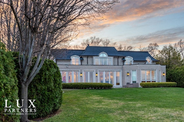 back of property at dusk featuring a high end roof, brick siding, a yard, and mansard roof