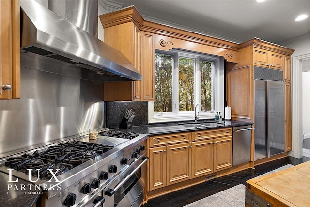 kitchen with backsplash, brown cabinets, wall chimney exhaust hood, premium appliances, and a sink