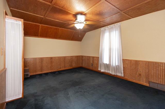 additional living space featuring wooden walls, dark colored carpet, a ceiling fan, and a wainscoted wall