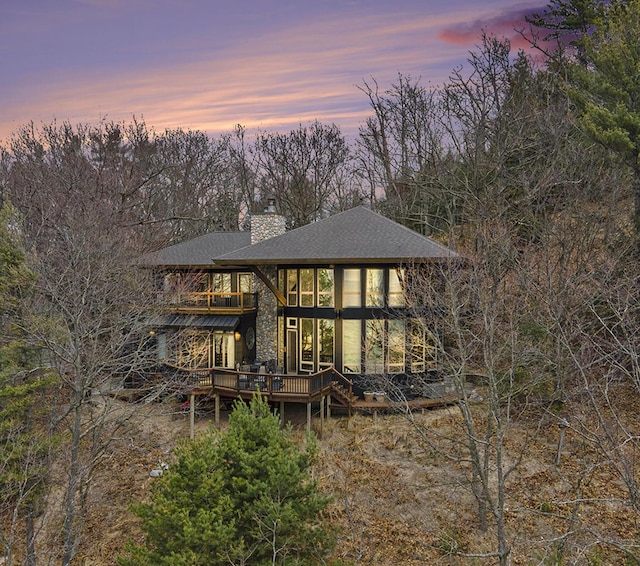 back of property at dusk with a chimney, a deck, a balcony, and a shingled roof