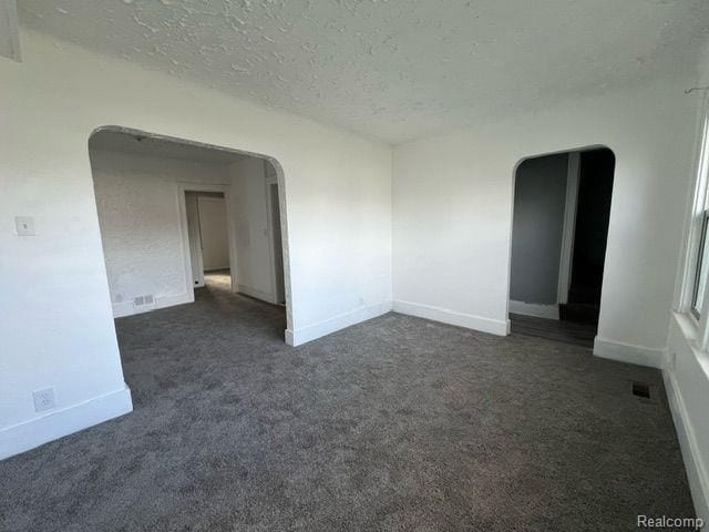 empty room featuring arched walkways, a textured ceiling, visible vents, baseboards, and dark carpet