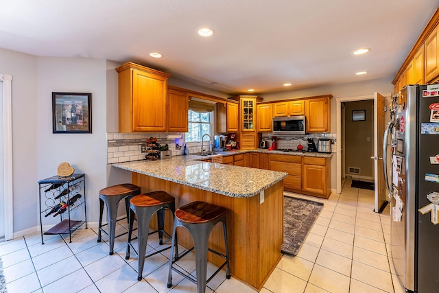 kitchen with a peninsula, tasteful backsplash, light stone counters, brown cabinetry, and appliances with stainless steel finishes