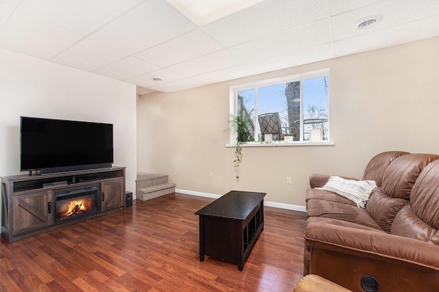 living area with a glass covered fireplace, a drop ceiling, dark wood finished floors, baseboards, and visible vents