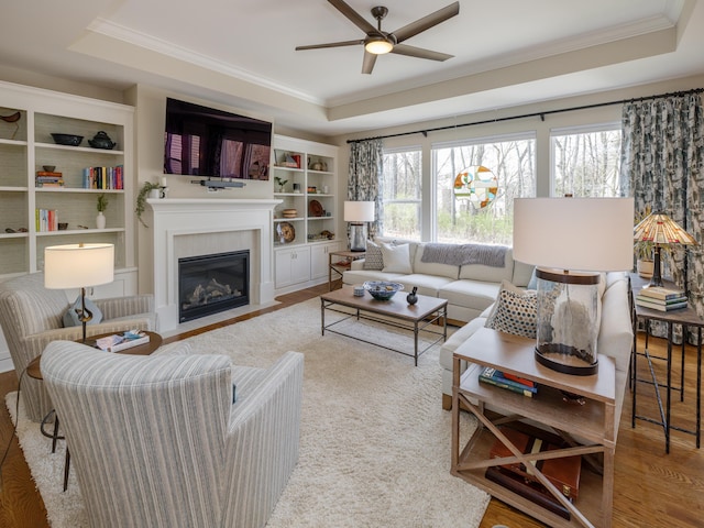 living room with a tray ceiling, a fireplace with flush hearth, ornamental molding, and ceiling fan