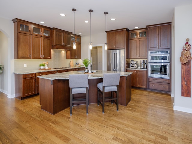 kitchen featuring arched walkways, a center island with sink, decorative backsplash, light wood-style flooring, and appliances with stainless steel finishes
