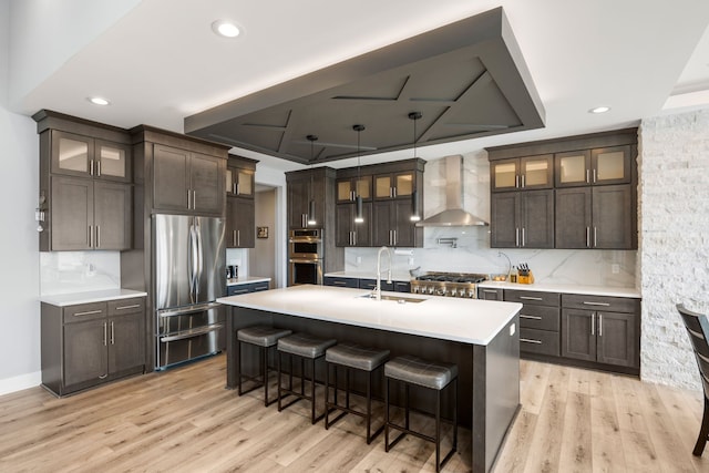 kitchen with wall chimney exhaust hood, a sink, dark brown cabinets, light wood-style floors, and appliances with stainless steel finishes