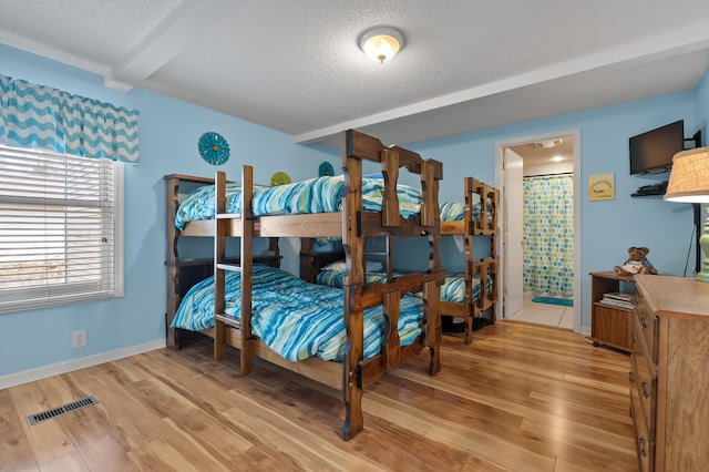 bedroom featuring a textured ceiling, light wood-type flooring, visible vents, baseboards, and beam ceiling