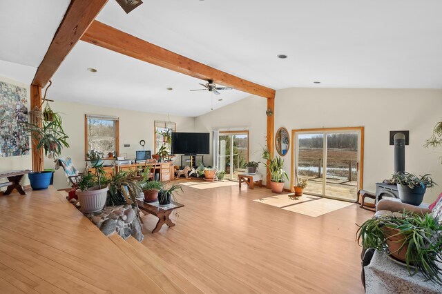 living room featuring wood finished floors, a wood stove, a ceiling fan, and vaulted ceiling with beams