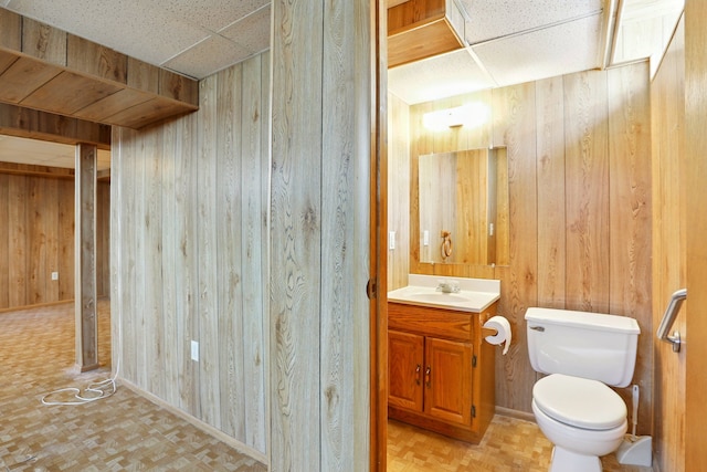half bath featuring wooden walls, a paneled ceiling, toilet, and vanity