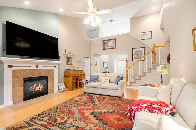 living room with stairway, wood finished floors, ceiling fan, a towering ceiling, and a tile fireplace