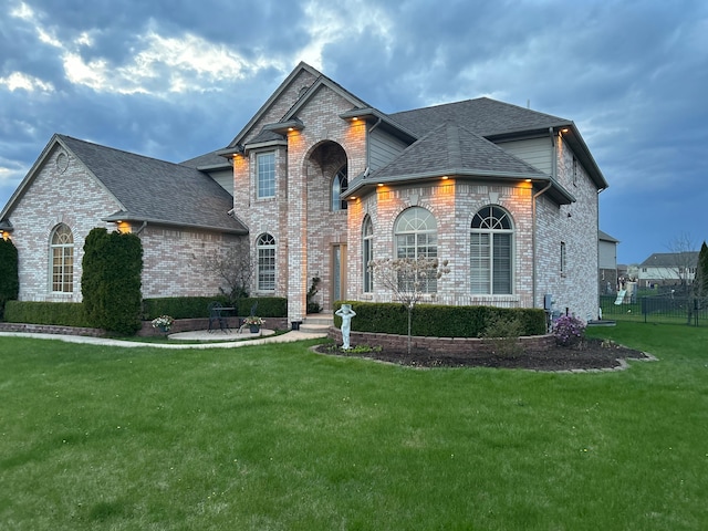 french country home featuring brick siding, roof with shingles, a front lawn, and fence