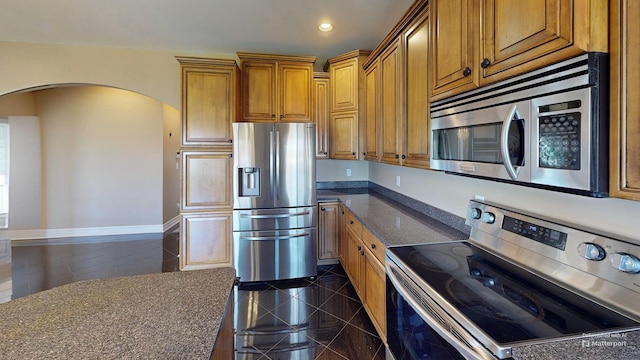 kitchen with recessed lighting, granite finish floor, arched walkways, brown cabinetry, and appliances with stainless steel finishes