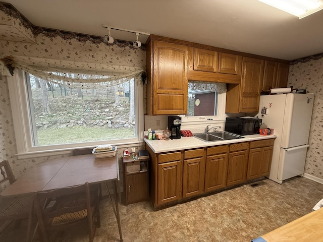kitchen with brown cabinets, wallpapered walls, a sink, freestanding refrigerator, and black microwave