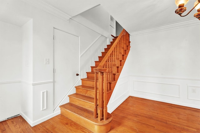 stairway with ornamental molding, wood finished floors, a notable chandelier, and a wainscoted wall