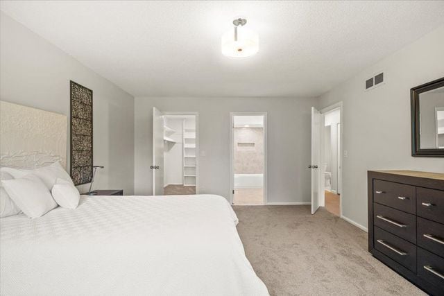 bedroom featuring visible vents, a walk in closet, ensuite bath, baseboards, and light colored carpet