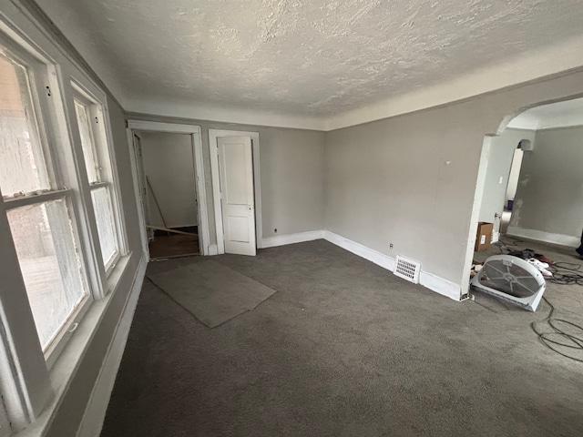 unfurnished bedroom featuring a textured ceiling, arched walkways, baseboards, dark colored carpet, and visible vents