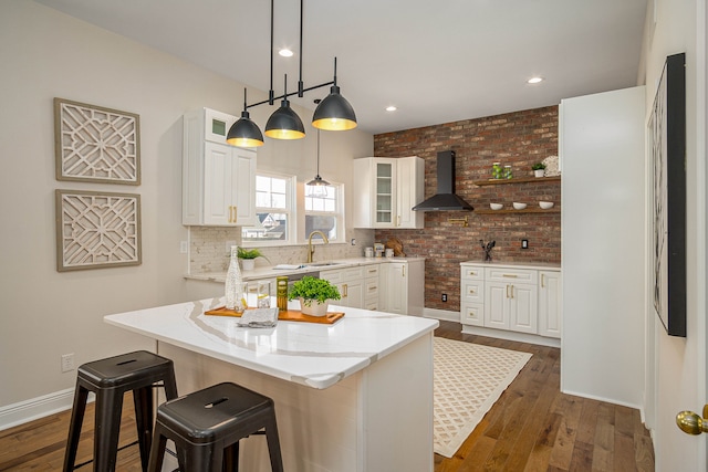 kitchen with wall chimney range hood, dark wood-type flooring, white cabinets, light stone counters, and glass insert cabinets