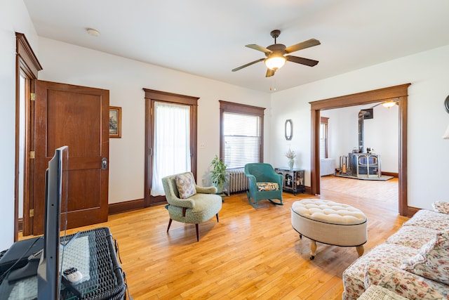 living room with light wood-style floors, radiator heating unit, ceiling fan, baseboards, and a wood stove
