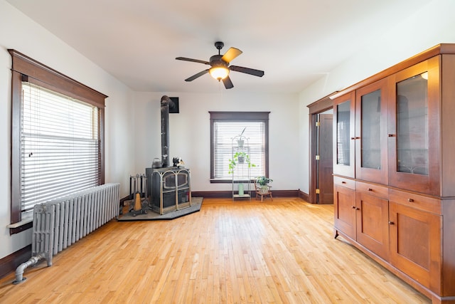 interior space featuring light wood-style flooring, a wood stove, ceiling fan, baseboards, and radiator