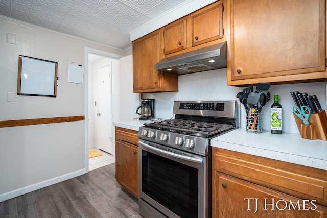 kitchen with under cabinet range hood, light countertops, stainless steel range with gas stovetop, dark wood-type flooring, and brown cabinetry
