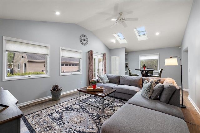 living room with ceiling fan, vaulted ceiling with skylight, baseboards, wood finished floors, and visible vents