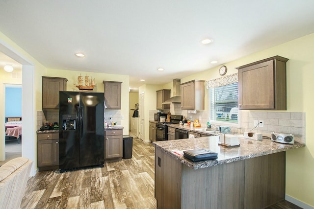 kitchen featuring light stone counters, wood finished floors, wall chimney exhaust hood, black appliances, and a peninsula