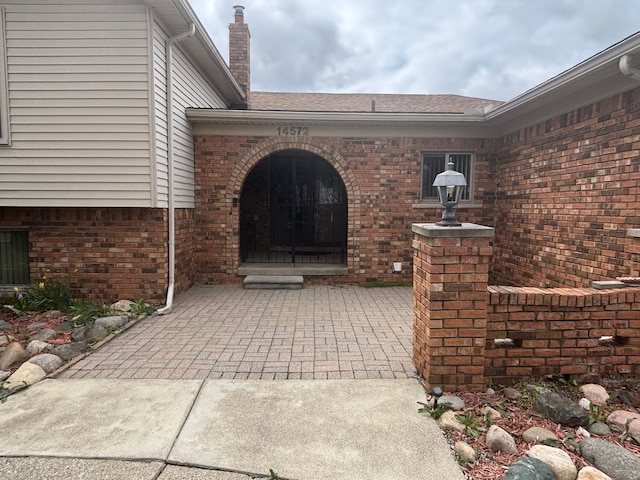 view of exterior entry with a chimney, brick siding, a shingled roof, and a patio area